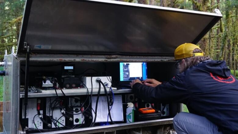 A man sits in the woods hunched over a computer screen placed on a steel work bench with numerous cords and electronic devices.