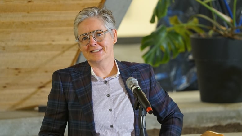 A woman with short grey hair and glasses stands at a microphone behind a podium.