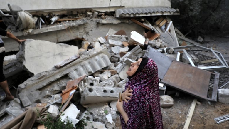 A woman in a hijab looks upward while standing in front of the concrete rubble of a destroyed building. 