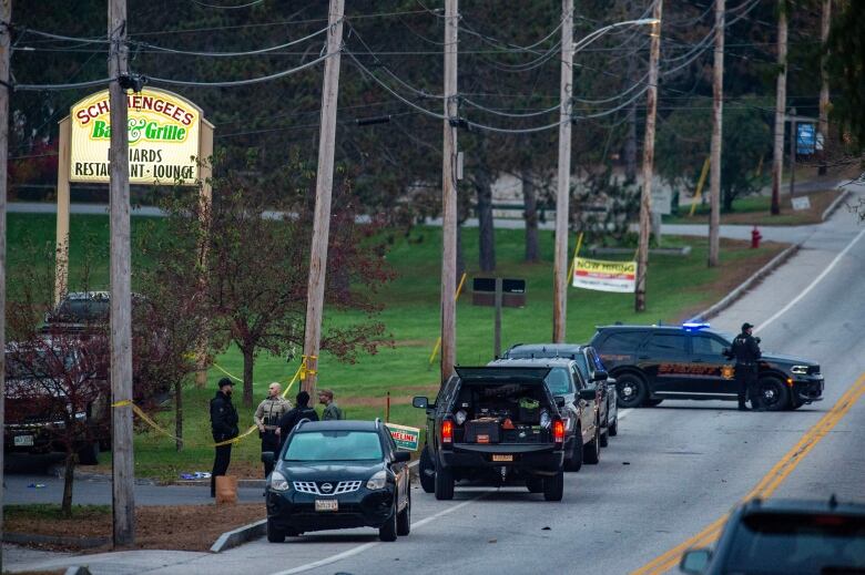 A photograph from a distance displays several cars and individuals, some wearing police uniforms, outside a business with a sign that reads 'Schemengees bar and grille: restaurant-billiards-lounge.'