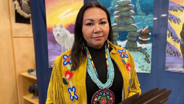 An Indigenous woman with mid-length, dark brown hair, is pictured smiling to the camera. She is wearing a beaded medallion and leather fringe jacket.