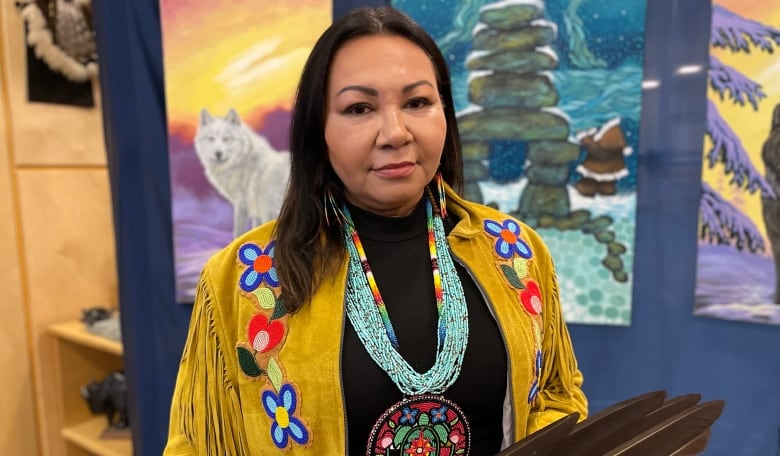 An Indigenous woman with mid-length, dark brown hair, is pictured smiling to the camera. She is wearing a beaded medallion and leather fringe jacket.