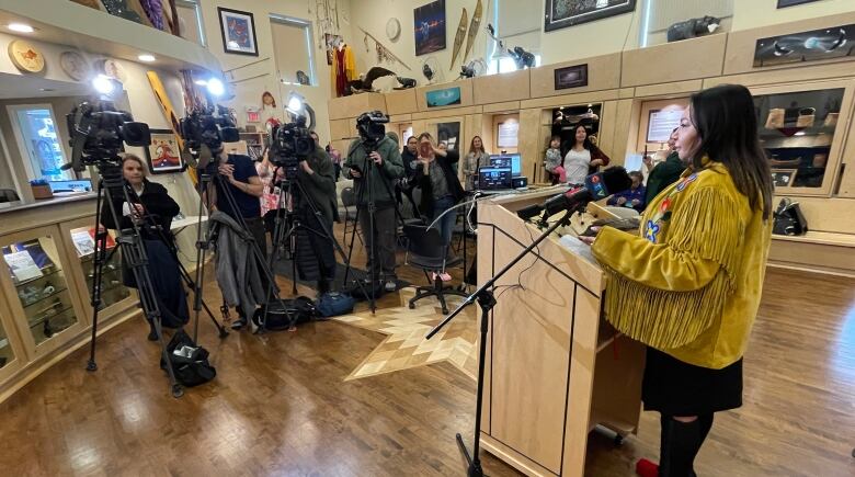 A woman is shown speaking at a podium, while four TV cameras and group of reporters in front of her.