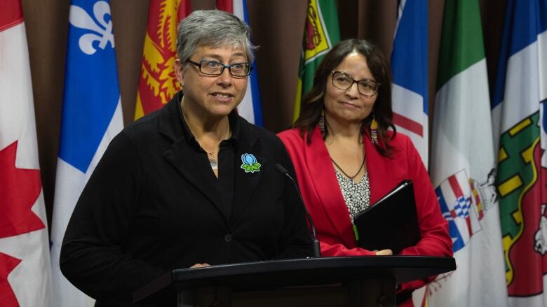 A politician speaks at a podium with flags behind her as another politician looks on.