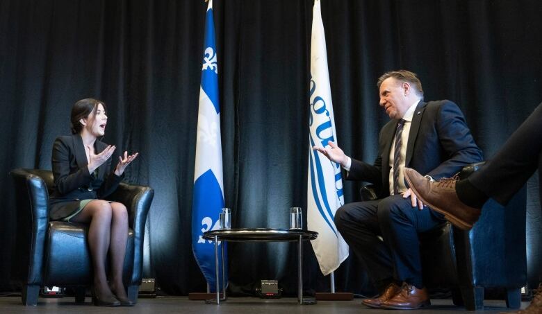 Woman and man sit in leather chairs on a stage. Behind them two flags hang. 