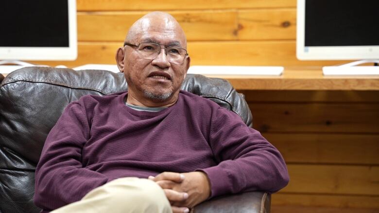 A man in a purple sweater and chino pants sits on a black couch. 