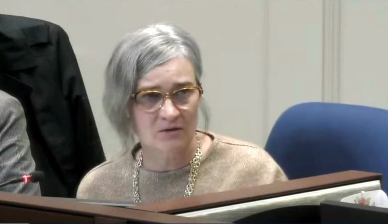 A white woman with gray hair pulled back behind her head, and glasses, sits at a desk with a microphone