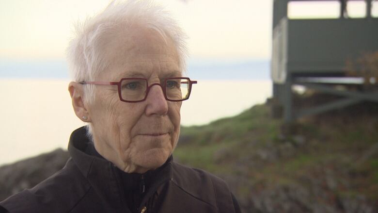 A woman with a black jacket and red-rimmed glasses stands in front of the ocean.