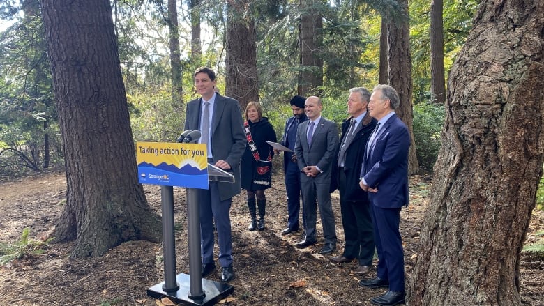 A man in a suit stands behind a lectern with a sign in yellow and blue that reads, 'Taking action for you,' and a logo of the B.C. provincial government on the bottom-left corner. He is outdoors, surrounded by trees, behind him four men in suits, including one wearing a turban, and a woman in a black outfit, with what looks like a sash featuring red and white blocks diagonally across her torso. 