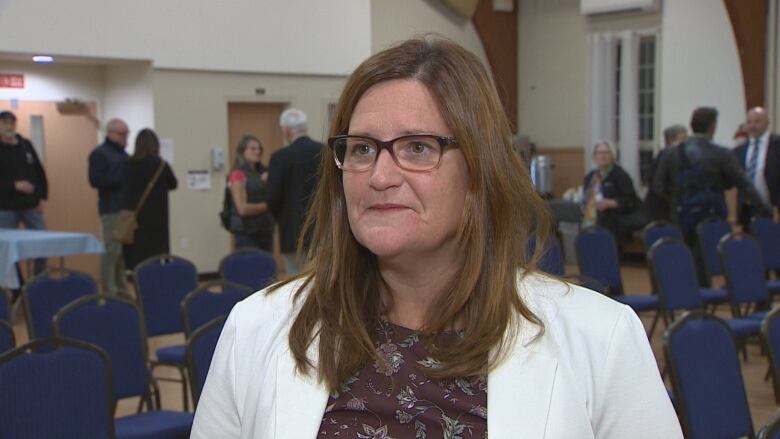 A woman with a white blazer, glasses and brown hair stands in a room during an interview.