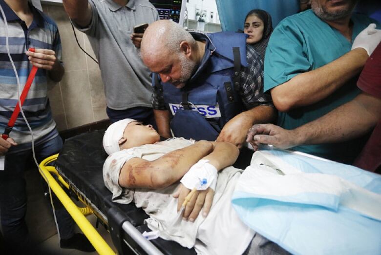 A man wearing a Press jacket stands over an injured child on a stretcher.