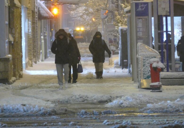 People walk along a snowy downtown street on a dark morning.