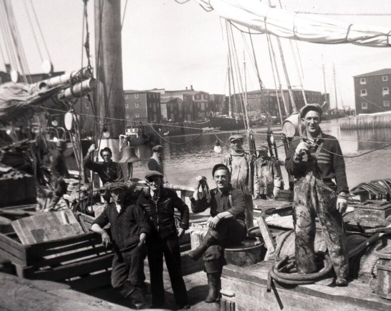 An archival image depicts six men sitting or standing on a wooden schooner docked in a harbour. 