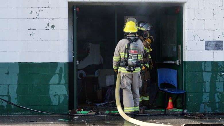 Two firefighters in full gear, including helmets, haul a hose into a green and white cinder-block building. There's smoke coming out the door and a large, white roller skate can be seen painted on the wall beside them.