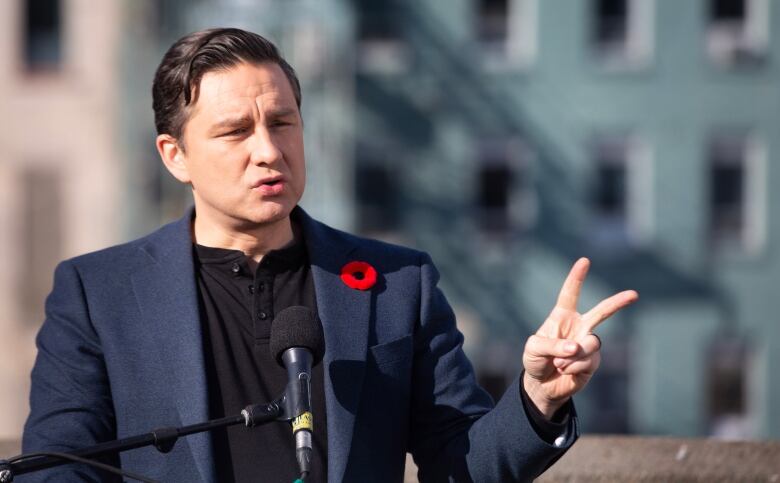 Pierre Poilievre, wearing a suit jacket over a t-shirt, speaks into a microphone. 