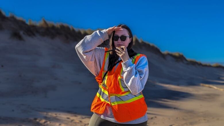 A woman in an orange safety vest talks into a radio on a beach with dunes in the background 