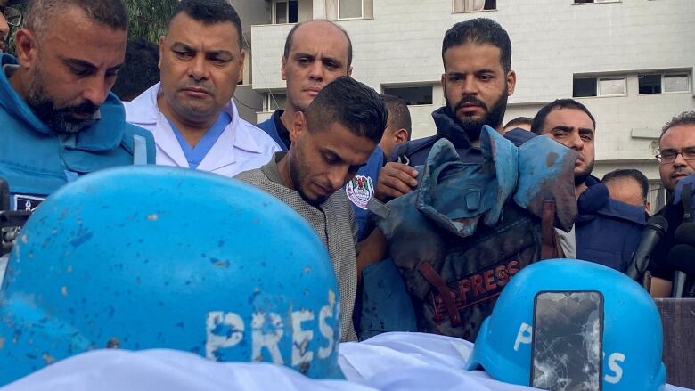 Helmets with the word, 'PRESS,' on them are seen displayed as people gather around.