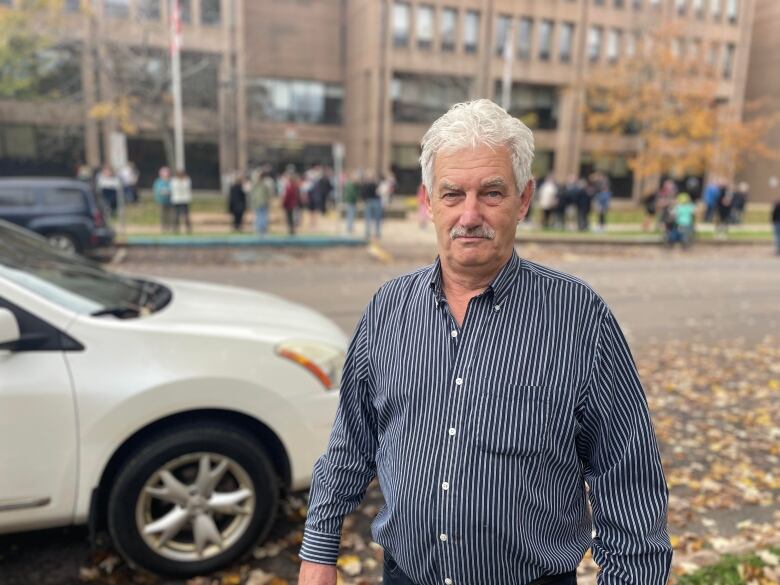 A man with grey hair and a mustache wears a blue stripped shirt and looks at the camera. A crowd of people gathers behild. 