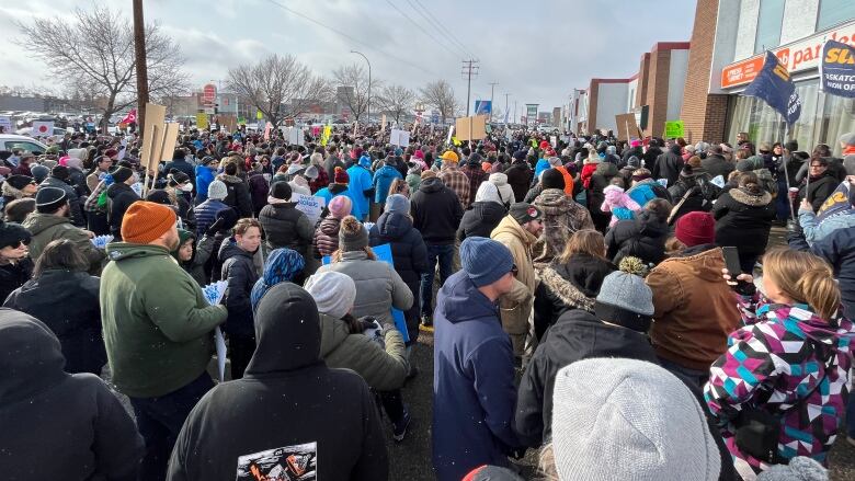 Hundreds of people are packed wearing winter clothing in a crowd outside a strip mall