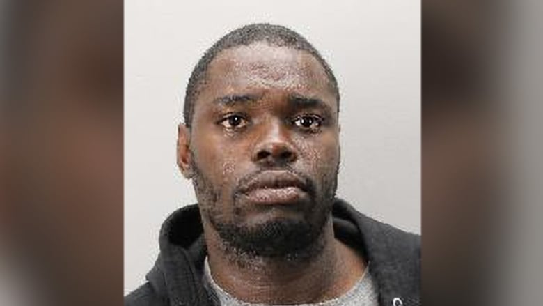 A headshot of a black man with brown eyes and short black hair. He is wearing a grey t shirt and dark grey hoodie.