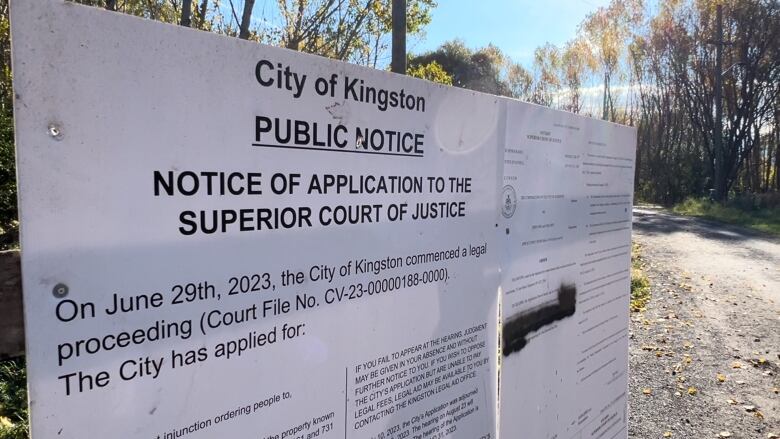 A legal document is printed out on two large, white boards. It explains Kingston is seeking an injunction to clear the park. It's a sunny, fall day and a gravel path and trees with yellow leaves can be seen in the background.