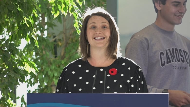 A woman with short, light brown hair wearing a black cardigan with white polka dots stands smiling at a podium.