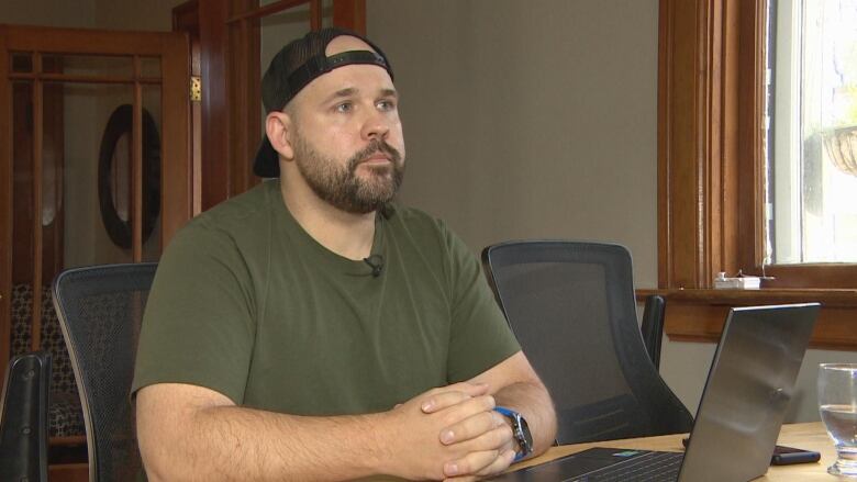 A man wearing a green t shirt and a black snap back can be seem sitting at a desk. 