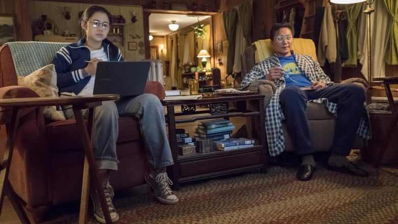 Leah Lewis (left) and Collin Chou (right) in The Half of It. They're sitting in chairs in their living room watching TV. 