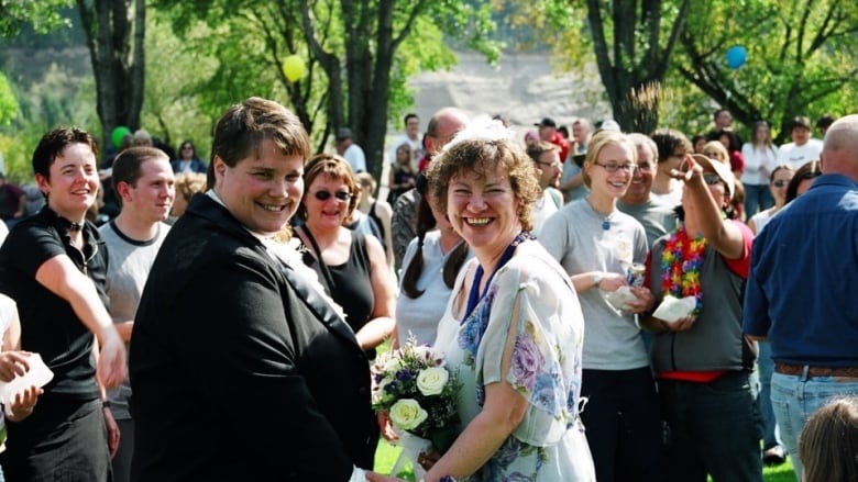 A beaming woman in a formal dress, with a bouquet, holds hands with a woman in a tuxedo as a crowd in a leafy park smiles and tosses confetti birdseed over the couple. 