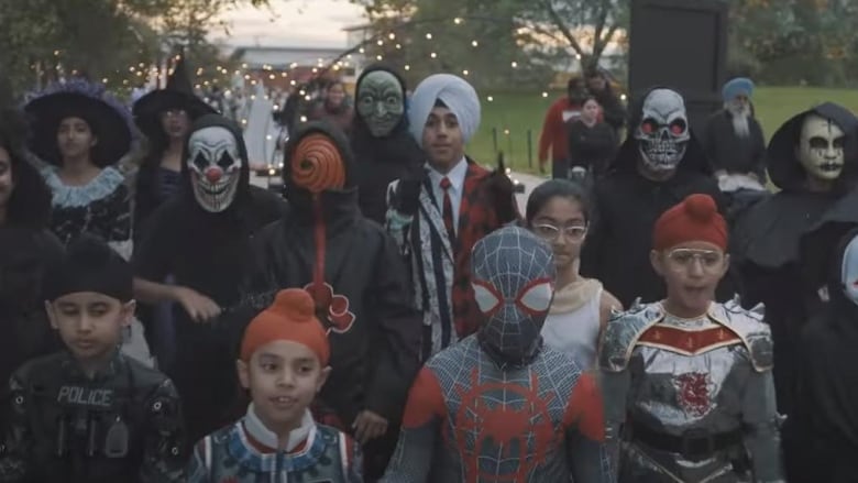 A young man in a turban stands in a crowd of costumed kids in a still from a music video. 