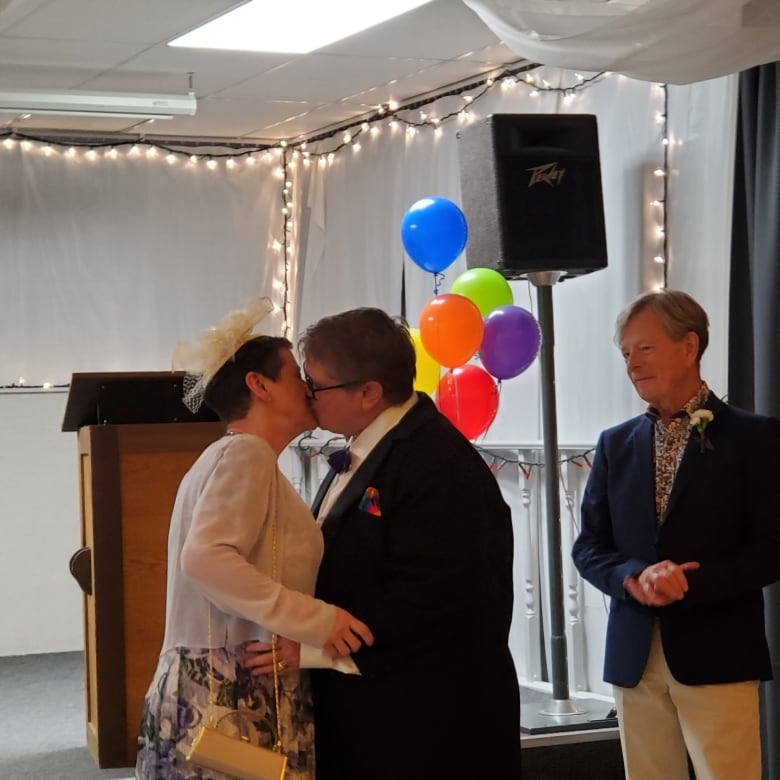 Two women in formal outfits kiss each other as a male wedding guest looks on. They're surrounded by colourful balloons and fairy lights. 