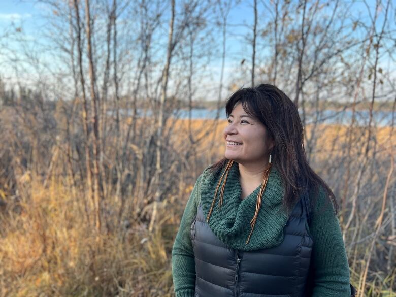 A woman wearing a sweater and a vest is standing in a patch of trees with a lake in the background. She is smiling and looking to the side. 