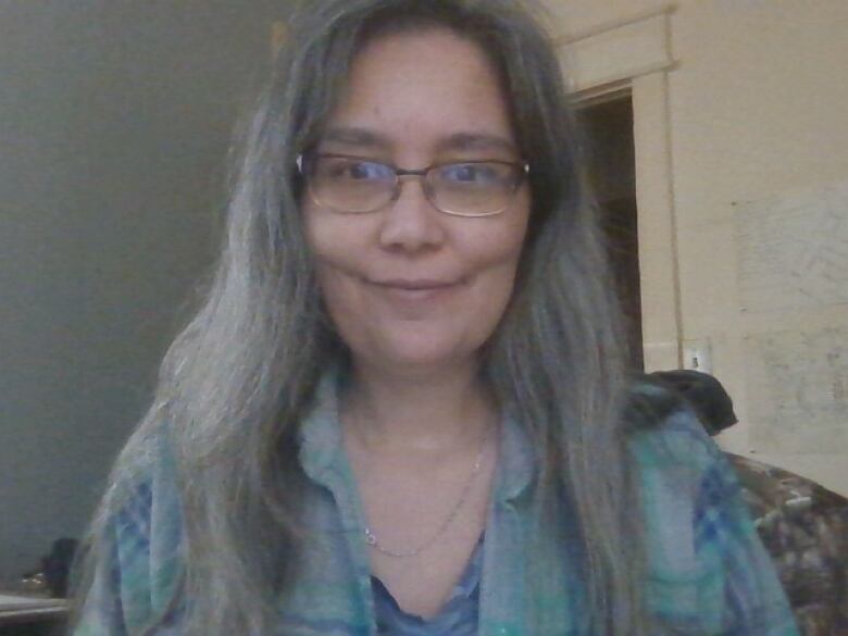 A woman indoors with long grey hair wearing rectangular glasses. 