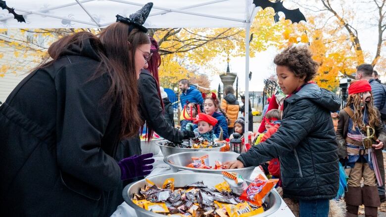 A child roots through a bowl of candy at an outdoor event. There's a line of costumed kids behind them.