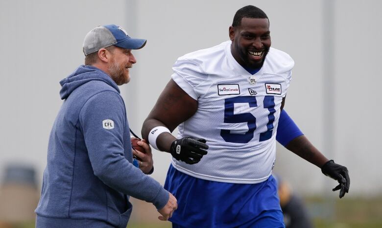 A man in a baseball cap, hooded sweatshirt and beard smiles and looks at a big football player in a white jersey, who is laughing.