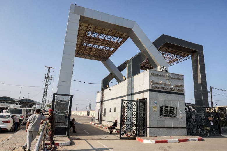 Men sit at the open gate of the Rafah border crossing, with cars on the left side and some pedestrians standing nearby.