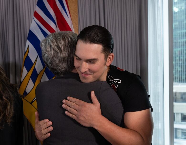 A man hugs a short-haired woman while a bald man looks on.