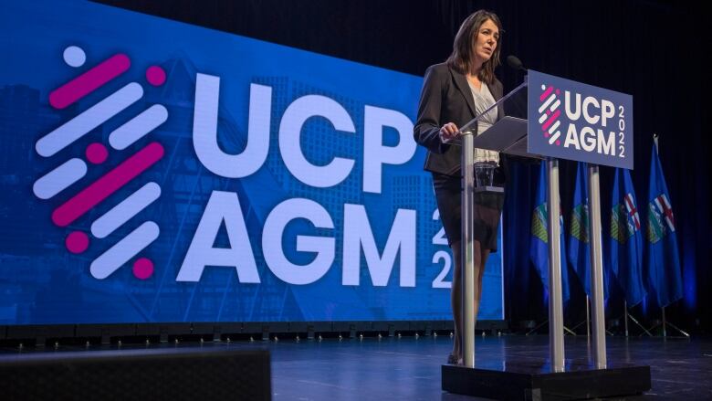 Alberta Premier Danielle Smith stands at a podium and speaks. Behind her is a screen that reads 