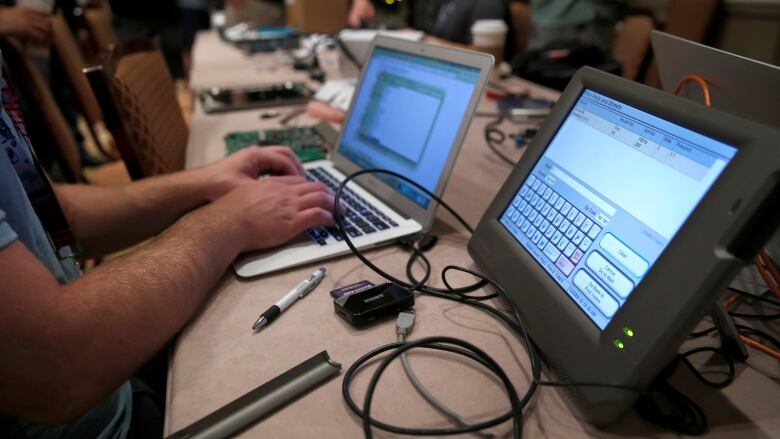 A person's hands are shown in front of multiple screens depicting computer hacking. 