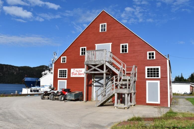 A red three-storey building has a wooden staircase leading to a door on the second floor. It sits behind a parking lot and in front of a body of water.