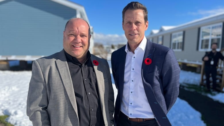 Two men in business jackets and open-throated shirts stand in front of two mini-homes. The ground is covered in snow but the sun is shining. 