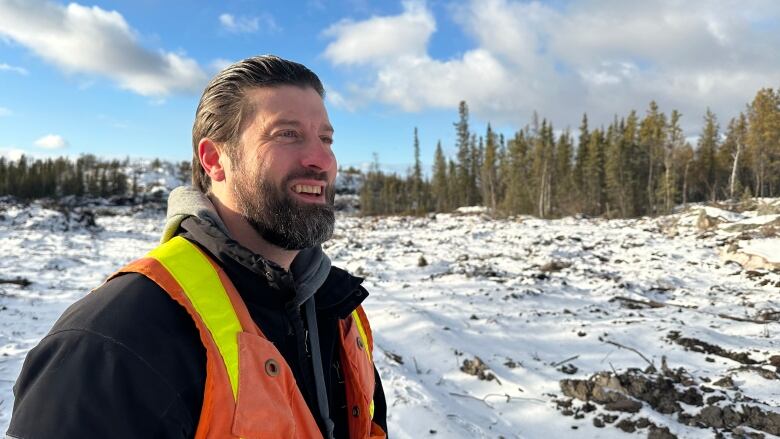 A man in a black coat with a safety vest on, stands outside. There's snow on rough ground around him, with trees in the background.