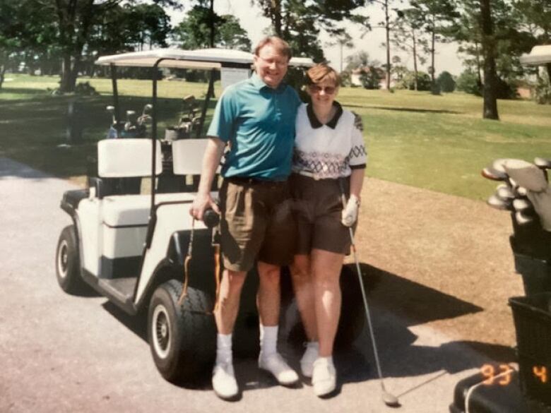 Man and woman in golf attire standing in front of golf cart. Woman holds a golf club.