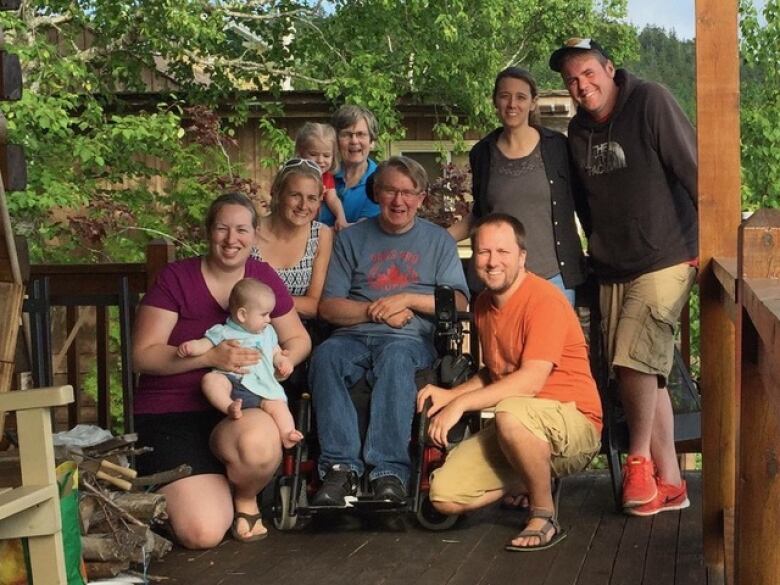 Man with grey hair and glasses sits in wheelchair surrounded by family including two small children.