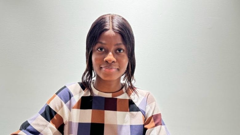 A dark haired woman wearing a patterned shirt sits at a table. The background is a plain white wall. She is looking at the camera with a slight smile on her face.