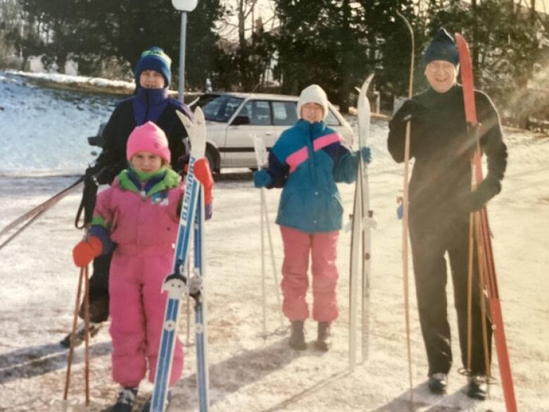 A man and three children dressed in ski wear stand in snowy area holding skis.