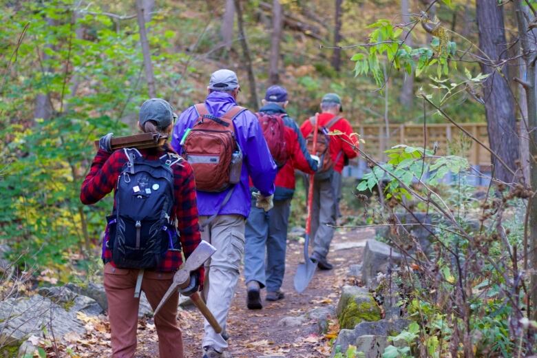 A group of four people carrying tools walk single file down a trail.