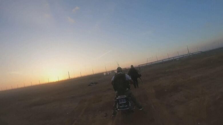 One man on a motorcycle and another on foot head toward a broken-down fence in dawn light.