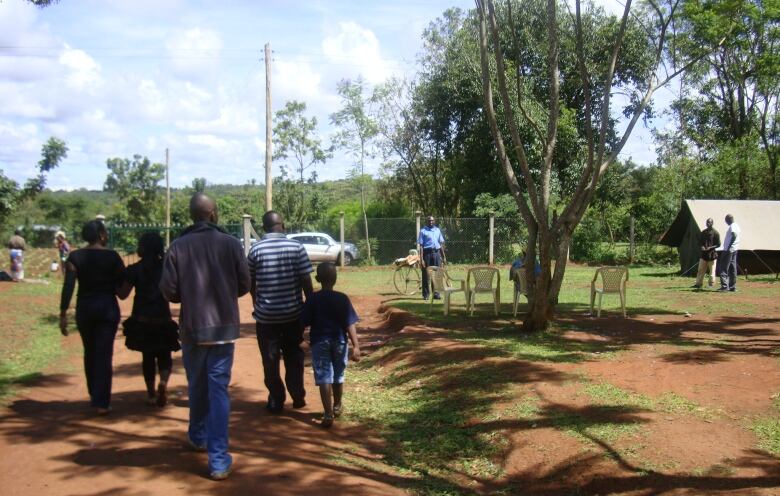 A small group of people, including two women, two men and a young boy, are seen from behind walking on a reddish dirt path. Around them are trees, some plastic outdoor chairs and other grownups talking or working.