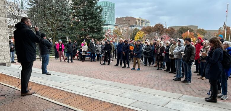 A man speaks into a microphone while people in winter jackets stand in a half circle around him